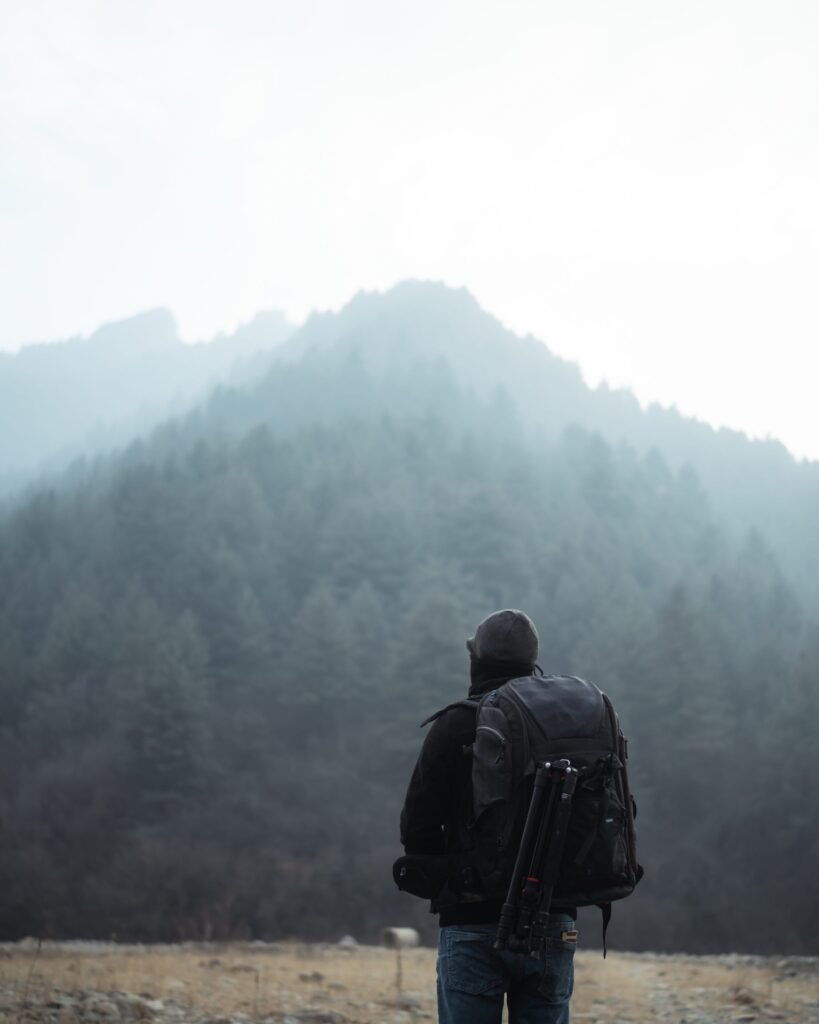 A person wearing a backpack looking at a large, forest covered mountain surrounded by mist. It gives a feeling of adventure and possibilities.