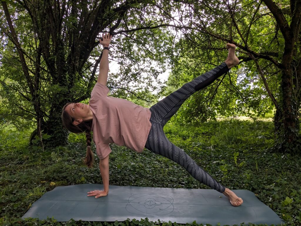 Yoga teacher in side plank outdoors, practicing vinyasa yoga under a large tree — promoting strength, balance and energy.
