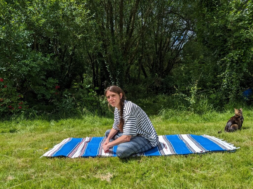 Yoga teacher in sitting on a blue stripy mat outdoors, practicing stillness in a sunny garden — finding mindfulness and peace.