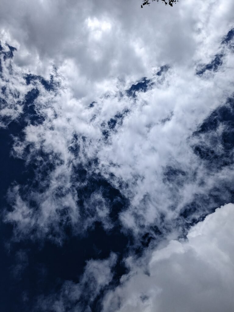 A photo of a deep blue sky, with large patchy white clouds with the sun behind them.